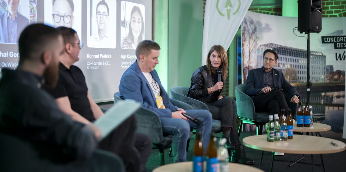 Na scenie odbywa się panel dyskusyjny z udziałem pięciu osób siedzących w nowoczesnych fotelach. Uczestnicy są zaangażowani, a kobieta w centrum trzyma mikrofon i zabiera głos. Za nimi widoczny jest duży ekran z portretami i nazwiskami panelistów, w tym „Konrad Weiske” i „Aga Kopytko”. Otoczenie jest profesjonalne i dobrze oświetlone, z zielonym światłem ambientowym oraz banerami Startup Wrocław i Concordia Design Wrocław w tle. Na małym okrągłym stoliku przed panelistami stoi kilka butelek wody i napojów. Atmosfera sugeruje formalne, lecz dynamiczne wydarzenie startupowe lub technologiczne, sprzyjające zaangażowaniu uczestników i wymianie wiedzy. Uczestnicy wydają się skupieni, uważnie słuchając lub przygotowując się do wypowiedzi, co podkreśla klimat współpracy i inspiracji.
