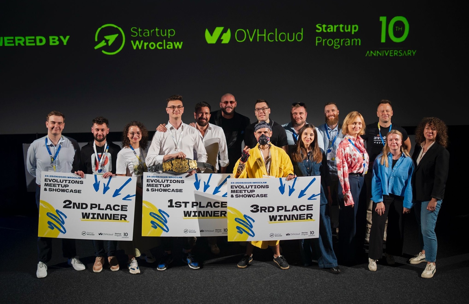 Grupowe zdjęcie uczestników i organizatorów wydarzenia „Evolutions Meetup & Showcase” we Wrocławiu. W centrum trójka laureatów trzymająca tablice z napisami: „1st Place Winner”, „2nd Place Winner”, „3rd Place Winner”. Za nimi ekran z logotypami: Startup Wrocław, OVHcloud i informacją o 10-leciu programu. Uczestnicy uśmiechnięci, w oficjalnych identyfikatorach.