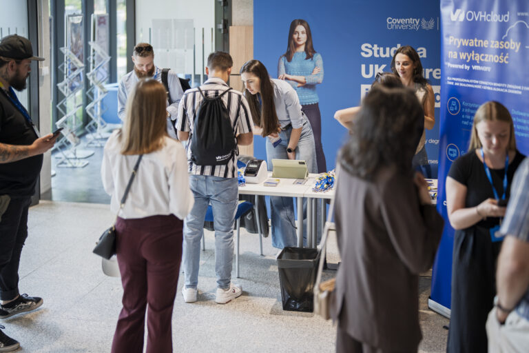 Strefa rejestracji tętni życiem. Uczestnicy wydarzenia zbierają się przy stoisku powitalnym, prowadzonym przez przedstawicieli Coventry University oraz OVHcloud. W tle widoczne są roll-upy informacyjne i branding partnerów. To moment pierwszych rozmów, wymiany uśmiechów, odbioru identyfikatorów i materiałów promocyjnych — networking w pełni.